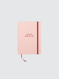 Rosa Hardcover-Notizbuch in minimalistischer Produktdarstellung auf hellem, neutralem Hintergrund. Auf dem Cover steht in roter Schrift „ES IST WIE ES IST“. Ein rotes Gummiband verläuft vertikal entlang der rechten Seite, dazu ein passendes rotes Lesebändchen unten. Das Notizbuch ist frontal ausgerichtet, symmetrisch positioniert und weich ausgeleuchtet.
