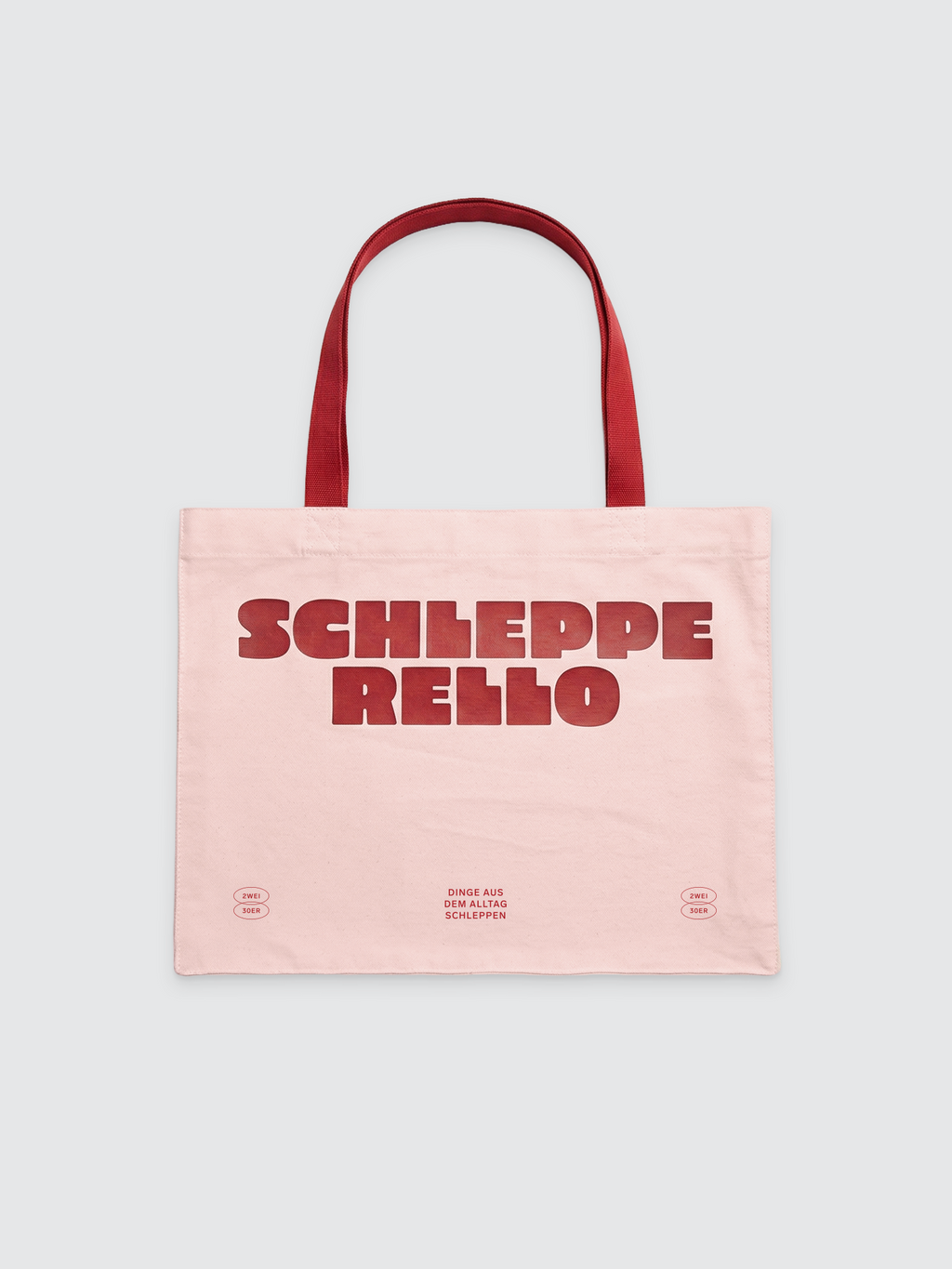 Eine hellrosa Canvas-Tote-Bag mit roten Henkeln. Auf der Vorderseite mittig ein großer, roter Schriftzug „SCHLEPPERELLO“ in einer fetten, leicht abgerundeten Blockschrift, der auf zwei Zeilen aufgeteilt ist. Am unteren Rand befinden sich drei kleinere rote Textelemente: In der Mitte steht „DINGE AUS DEM ALLTAG SCHLEPPEN“, flankiert von zwei kleinen ovalen Logos mit der Aufschrift „2WEI 30ER“.