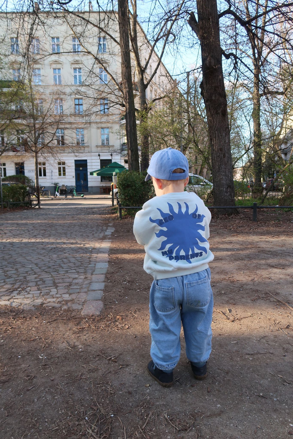Kleinkind steht mit dem Rücken zur Kamera auf einem Parkweg und trägt einen hellen Pullover mit blauem Sonnenmotiv und „CEO of“-Schriftzug.