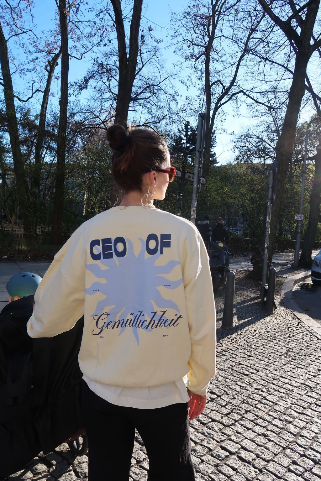 Person steht mit dem Rücken zur Kamera auf einem sonnigen Gehweg im Park und trägt einen cremefarbenen Pullover mit großem Sonnenmotiv und dem Schriftzug „CEO of Gemütlichkeit“.