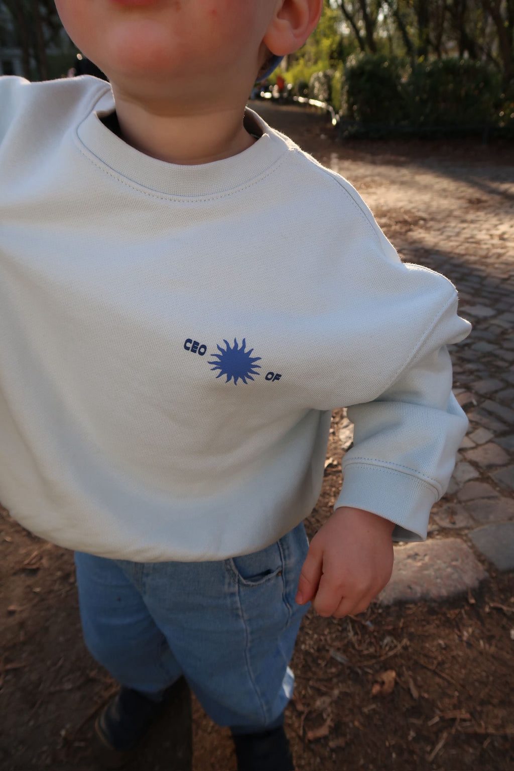 Nahaufnahme eines Kinderpullovers in hellem Ton mit kleinem „CEO of“-Print und blauem Sonnenmotiv auf der Brust, im Hintergrund ein sonniger Parkweg.