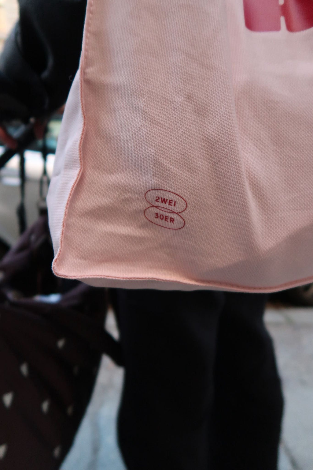 Nahaufnahme einer hellen Stofftasche mit kleinem „ZWEI 30ER“-Print in Rot, im Hintergrund unscharfe Straßenszene.