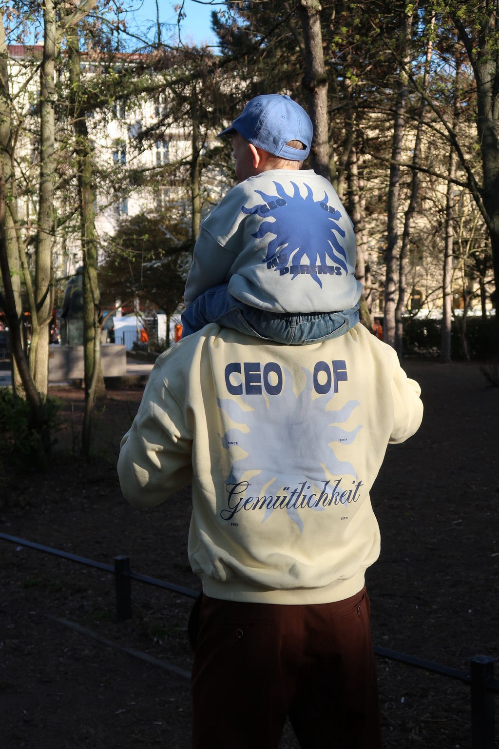 Erwachsene Person trägt ein Kind auf den Schultern im Park; beide tragen helle Oberteile mit großem Sonnenmotiv und „CEO of Gemütlichkeit“-Schriftzug auf dem Rücken.