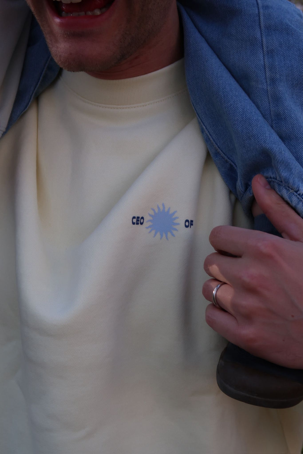 Nahaufnahme eines cremefarbenen Pullovers mit kleinem „CEO of“-Print und Sonnenmotiv auf der Brust; eine Hand hält eine blaue Jacke über der Schulter.