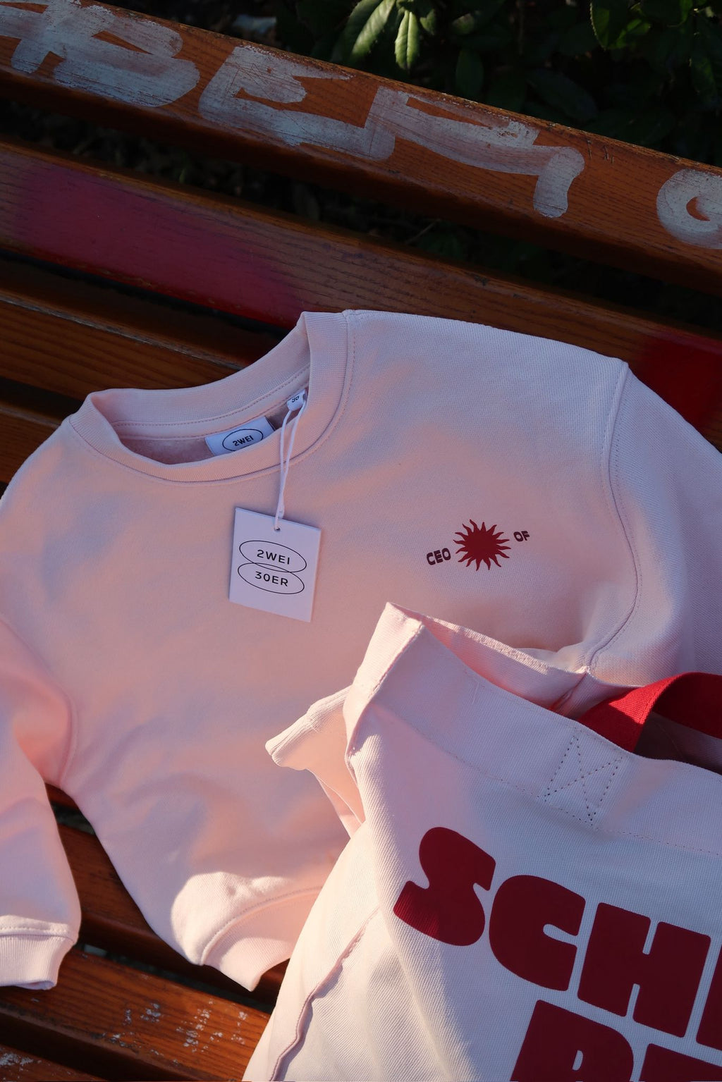 Sweatshirt und Tasche liegen auf einer Holzbank; Sweatshirt zeigt ein kleines „CEO of“-Logo auf der Brust, Tsche einen großen roten Schriftprint.