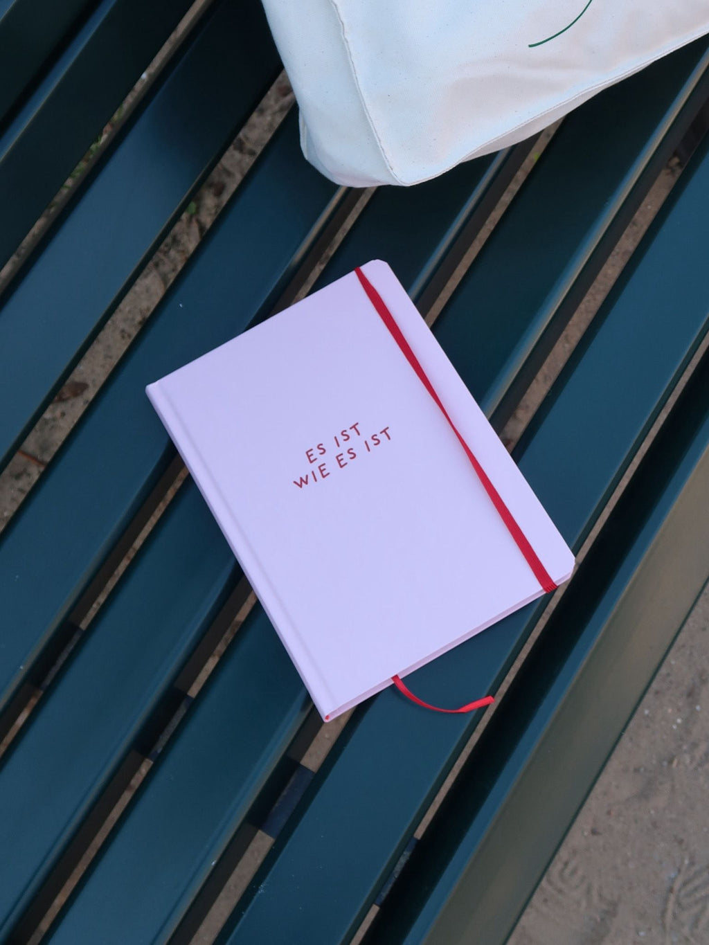 Geschlossenes rosa Notizbuch mit rotem Gummiband liegt auf einer grünen Parkbank; auf dem Cover steht „ES IST WIE ES IST“.