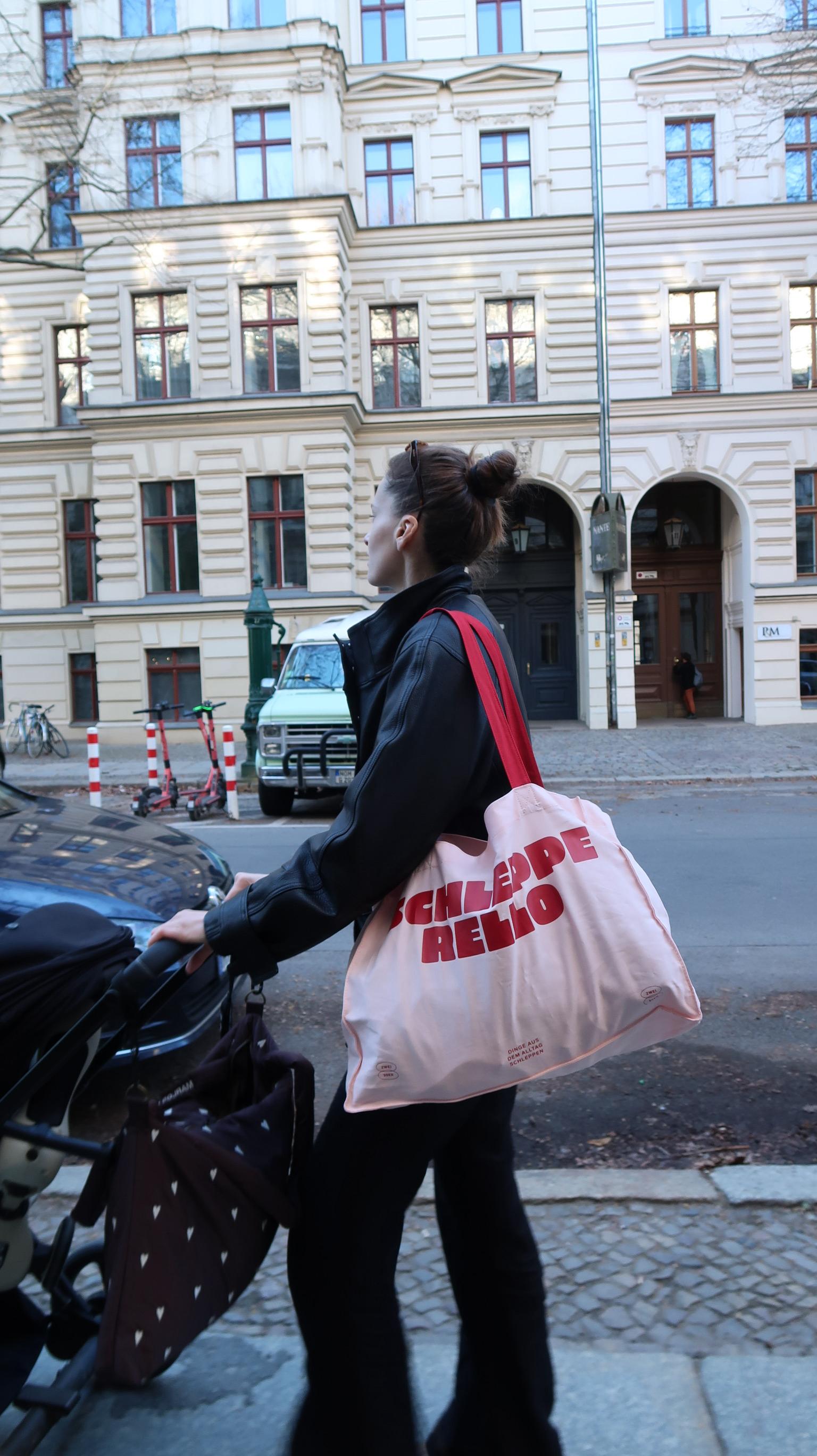 Person überquert eine Straße und trägt eine große helle Stofftasche mit rotem Schriftprint über der Schulter; im Hintergrund Stadtfassade.