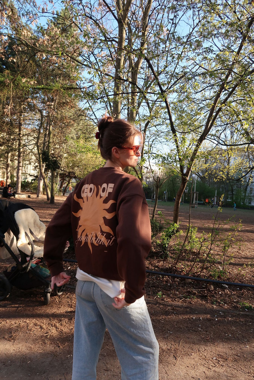 Person steht in einem sonnigen Park und trägt denselben braunen Pullover mit großem Sonnenmotiv und Schriftzug „CEO of …“ auf dem Rücken; umgeben von Bäumen und Frühlingslicht.