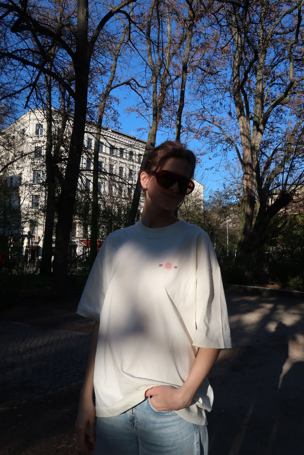Person mit Sonnenbrille steht im Park und trägt ein weißes T-Shirt mit kleinem „CEO of“-Print auf der Brust; warmes Sonnenlicht und Bäume im Hintergrund.