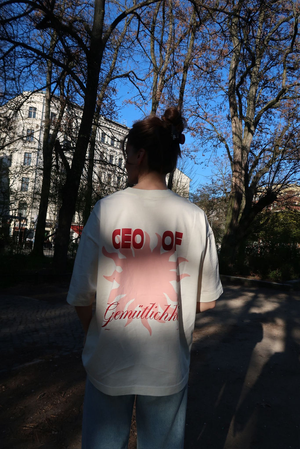 Person steht mit dem Rücken zur Kamera in einem sonnigen Park und trägt ein weißes T-Shirt mit großem roten Sonnenmotiv und dem Schriftzug „CEO of Gemütlichkeit“.