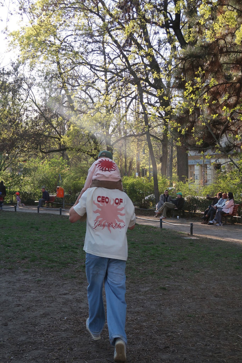 Erwachsene Person läuft durch einen Park mit einem Kind auf den Schultern; beide tragen passende Shirts mit rotem Sonnenmotiv und „CEO of“-Print auf dem Rücken, im Hintergrund sitzen Menschen auf Bänken.