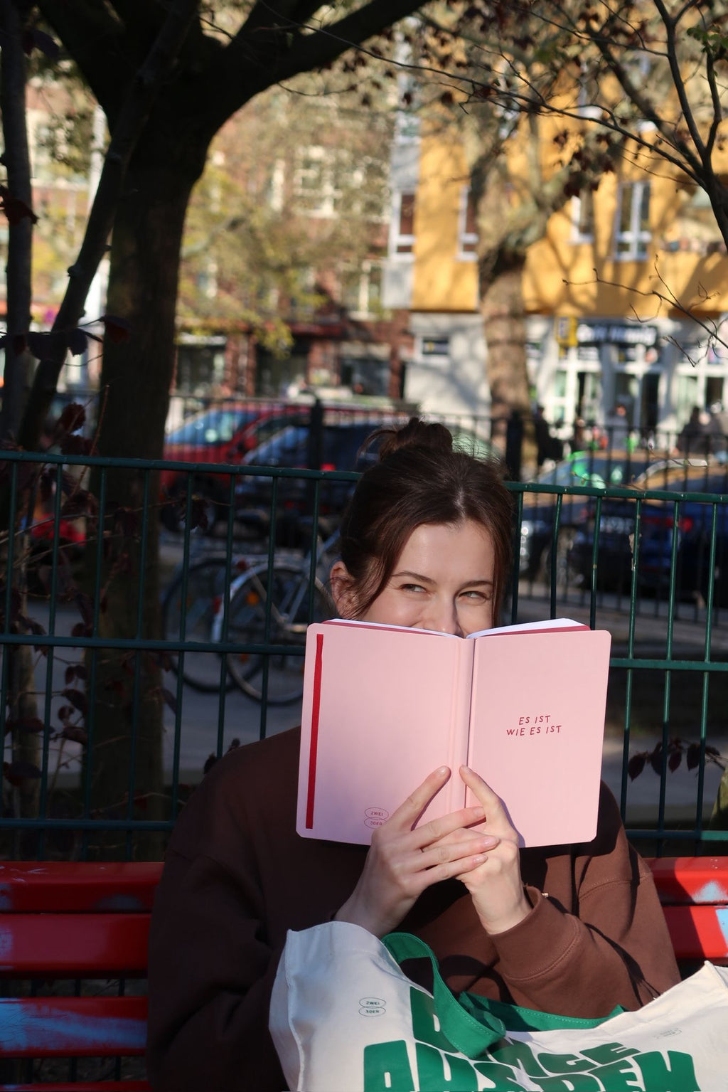 Person sitzt auf einer roten Parkbank und hält ein rosa Notizbuch mit der Aufschrift „ES IST WIE ES IST“ vor das Gesicht; im Hintergrund eine Straße mit Bäumen und Gebäuden.