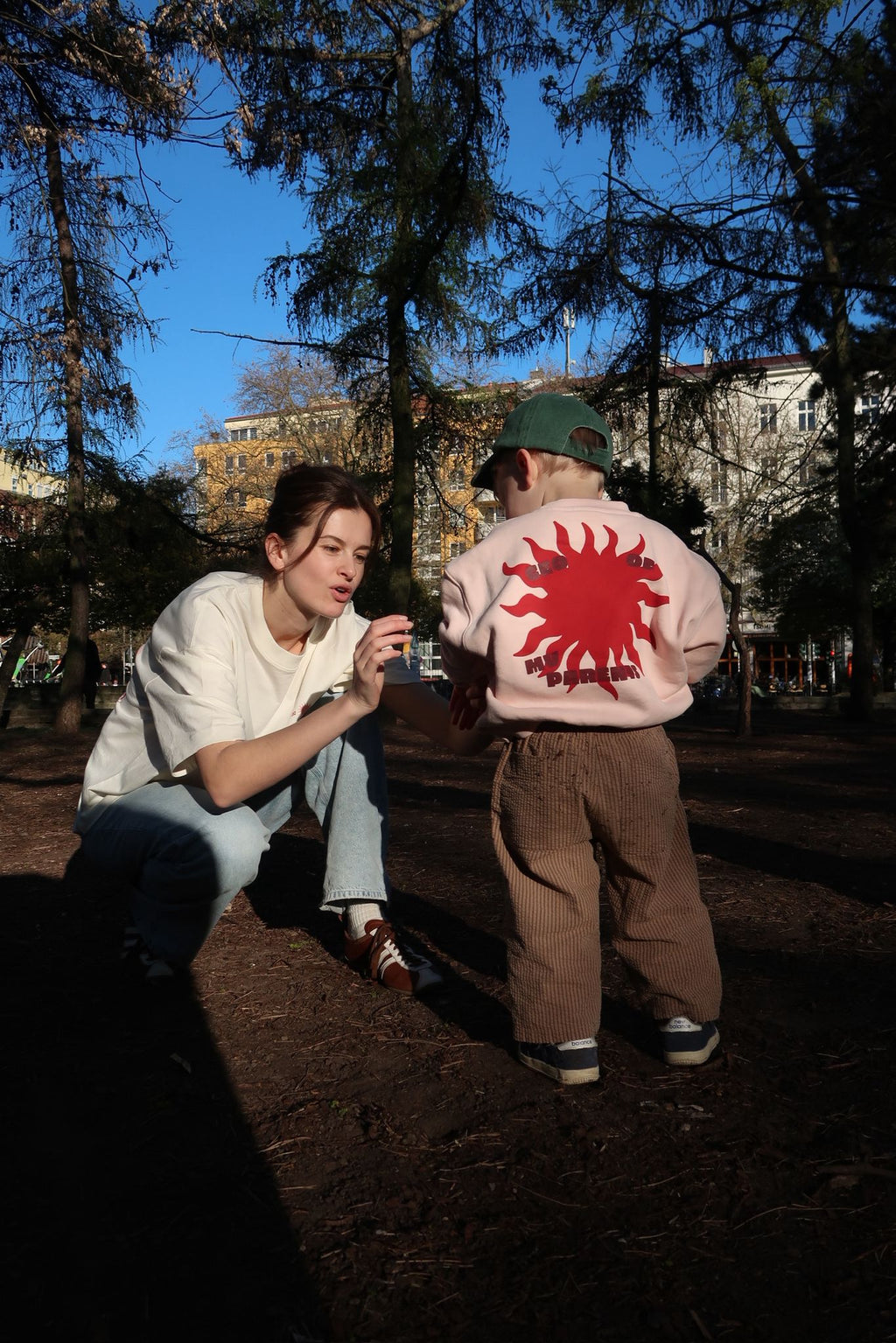 Person kniet im Park und spricht mit einem Kind, das einen rosa Pullover mit großem roten Sonnenmotiv und „CEO of“-Print trägt; warmes Abendlicht zwischen den Bäumen.